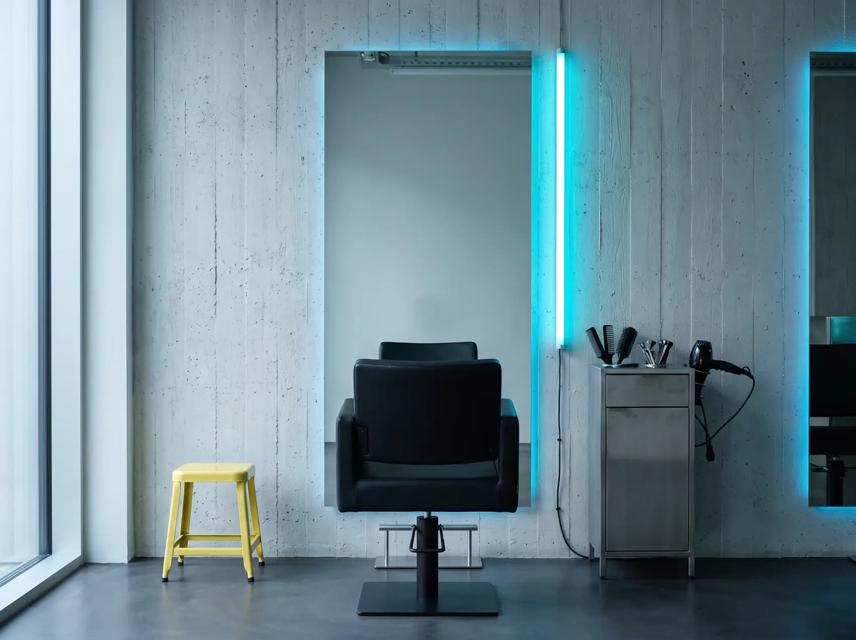 Ein einzelner Friseurplatz: schwarzer Stuhl vor einem großen Spiegel, daneben eine Neonröhre und ein Wagen mit Kämmen, Klammern und Föhn. Symbolbild.
